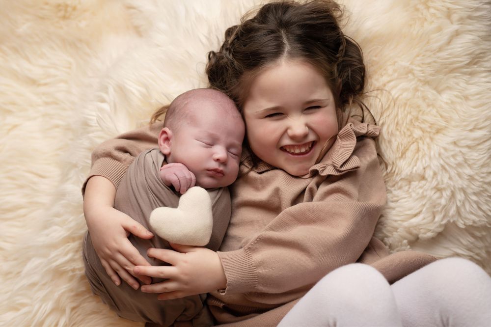 A smiling young girl lovingly holds a sleeping newborn baby wrapped in a beige swaddle on a soft fur blanket.