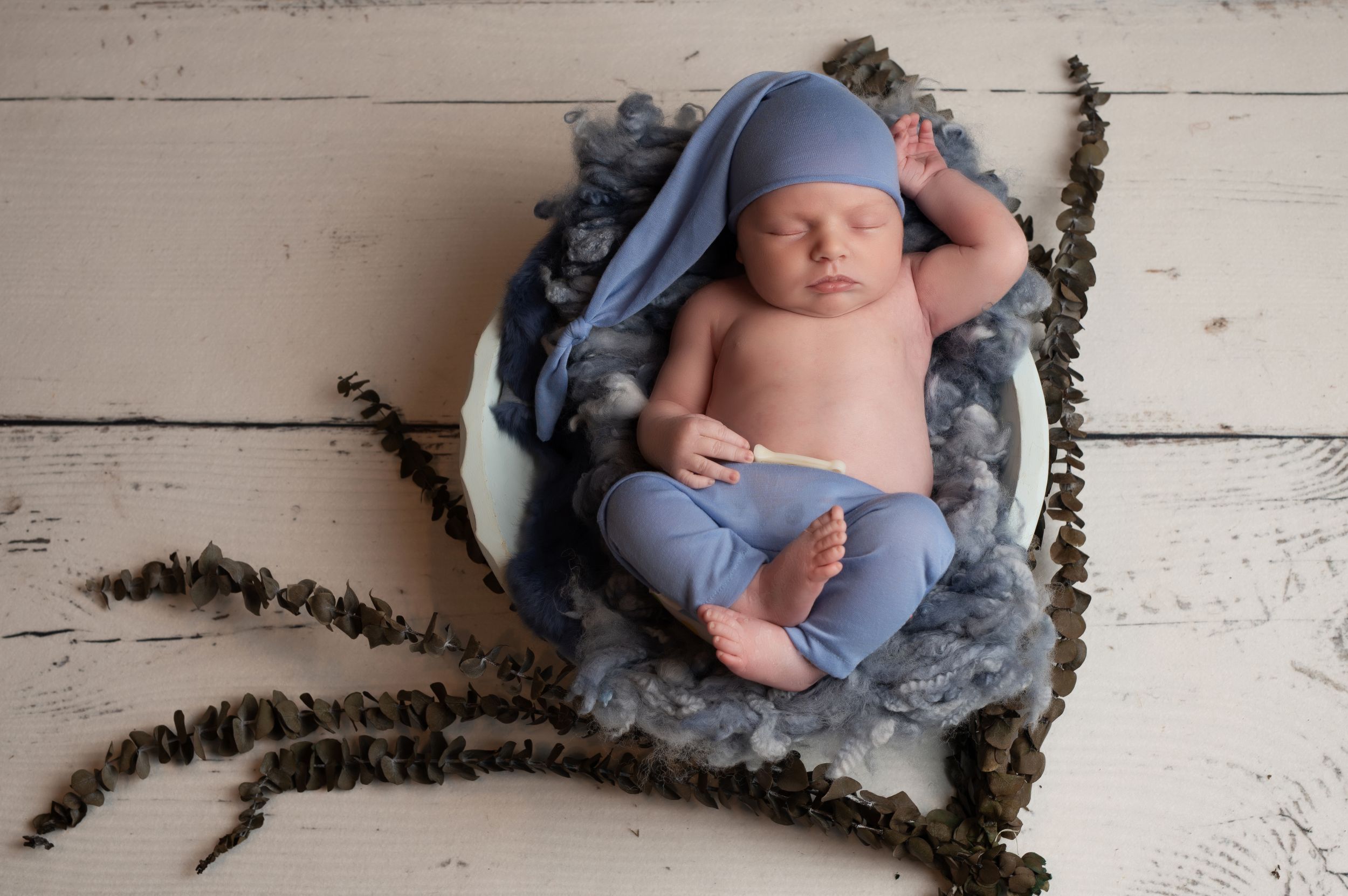Sleeping newborn baby in blue hat and pants nestled in a white bowl with soft blue fur on a rustic white wood floor.