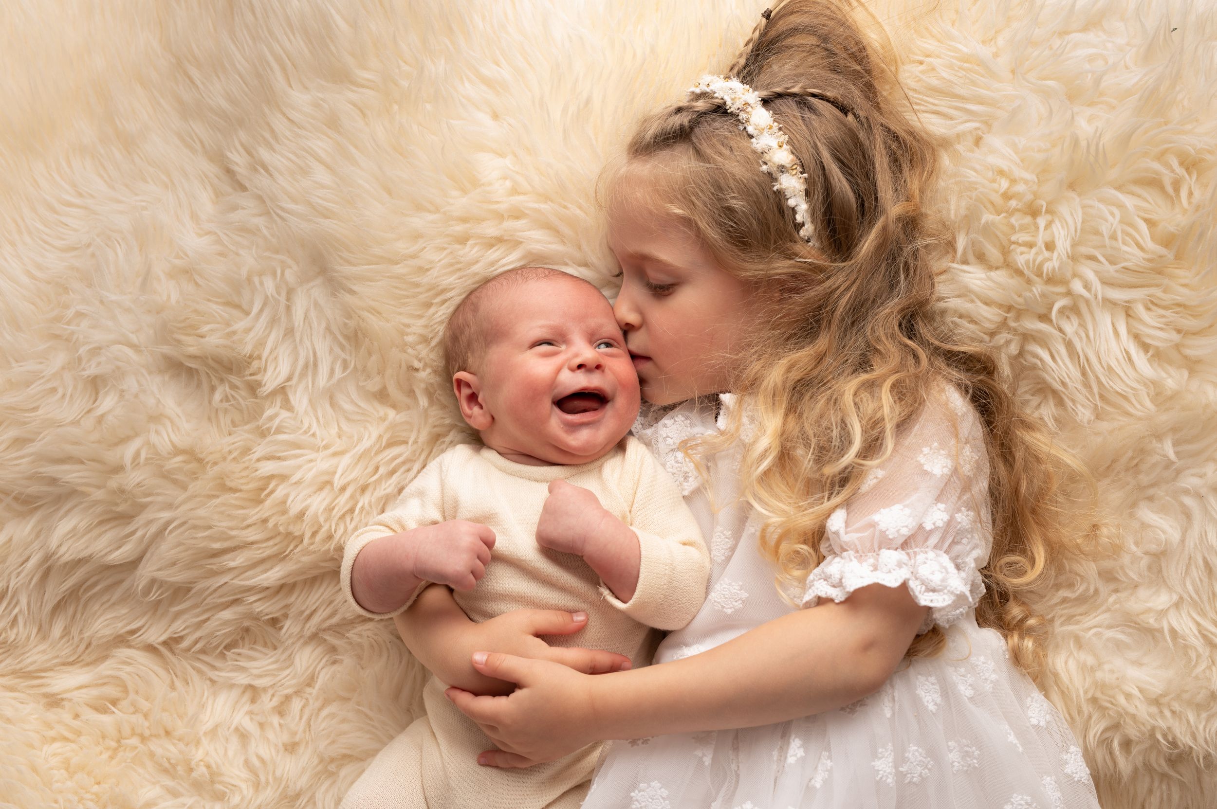 A young girl in a white dress kisses a laughing newborn baby on a soft cream fur blanket.