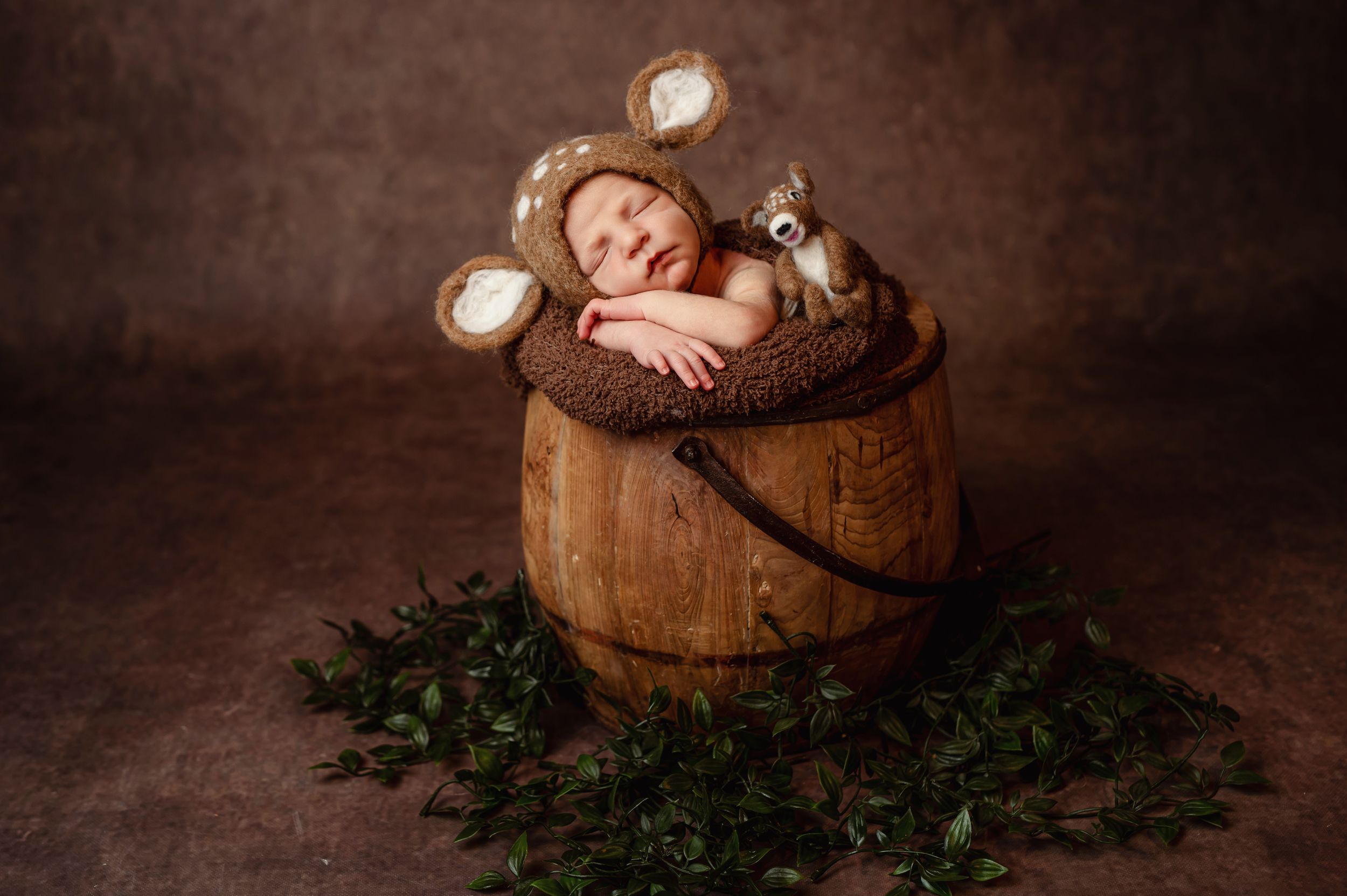 Sleeping baby in mouse ears prop nestled inside rustic wooden barrel surrounded by greenery on brown backdrop.