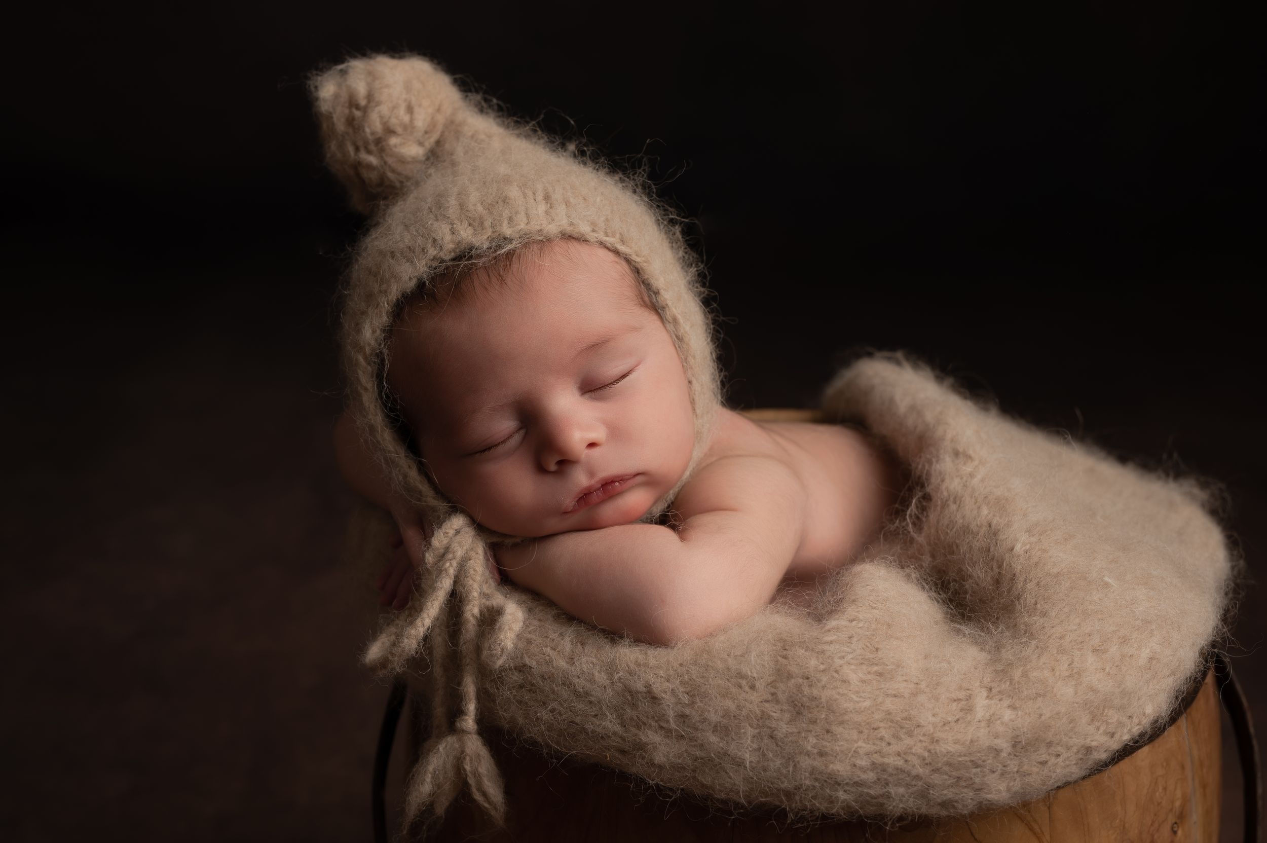 Sleeping newborn baby in a wooden bucket wearing a cozy beige knit bonnet hat, wrapped in soft wool blanket.