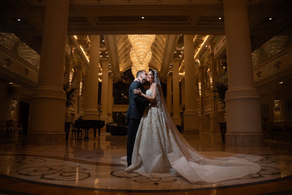 Bride and groom share a romantic kiss in an elegant grand ballroom with ornate chandeliers and marble floors.