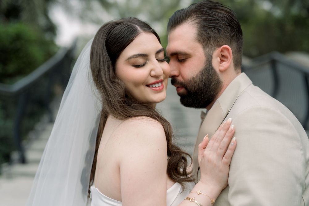 Romantic wedding portrait of a couple embracing outdoors, bride in white dress with veil smiling with groom in beige suit.