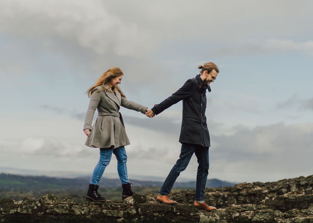 Two figures walking hand in hand along a rocky hilltop against a cloudy sky.
