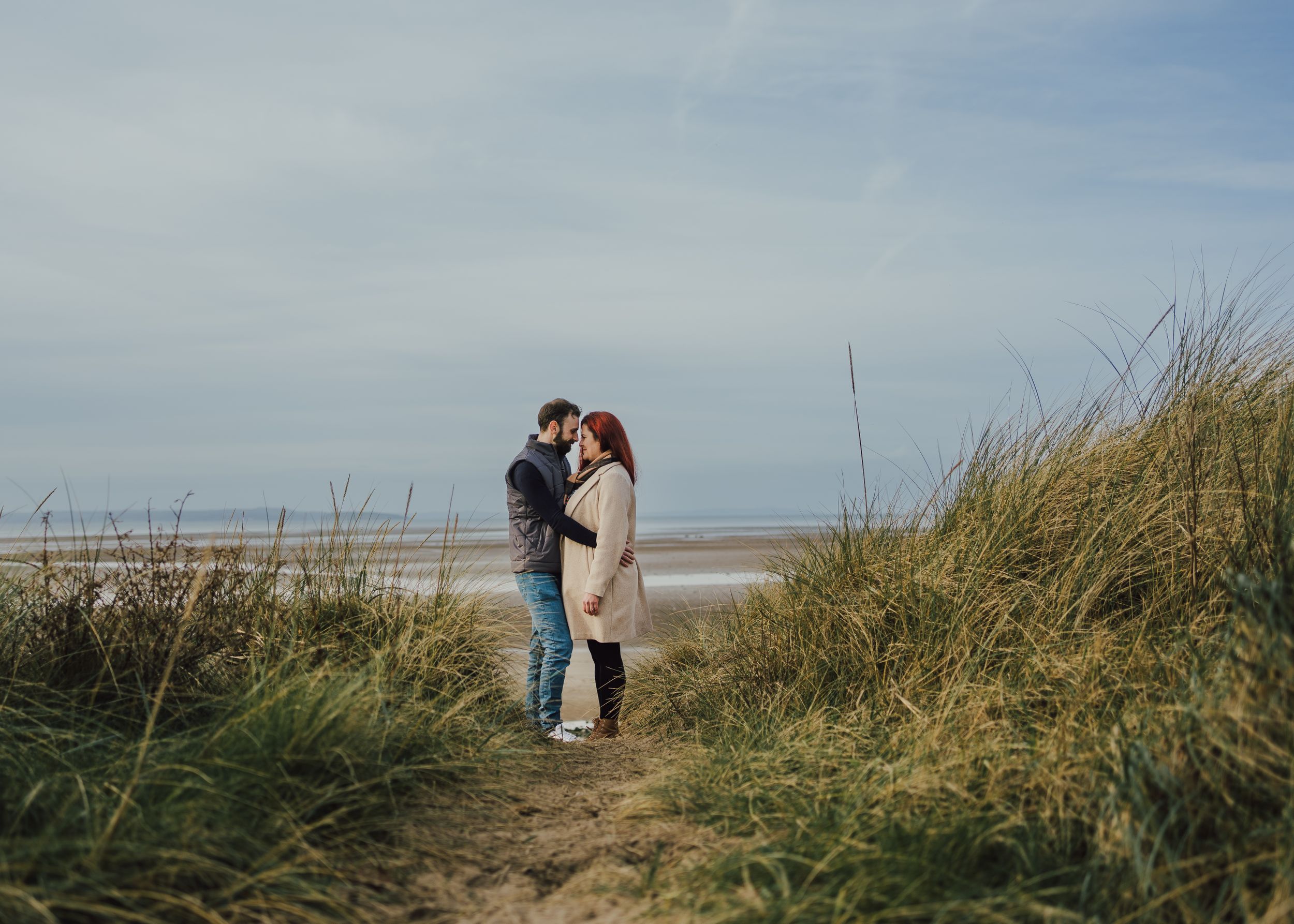 A couple embraces on a coastal path among tall beach grass with the ocean and cloudy sky in the background.