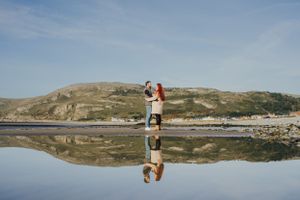 Two people embrace on reflective water surface against rolling hills and blue sky landscape.