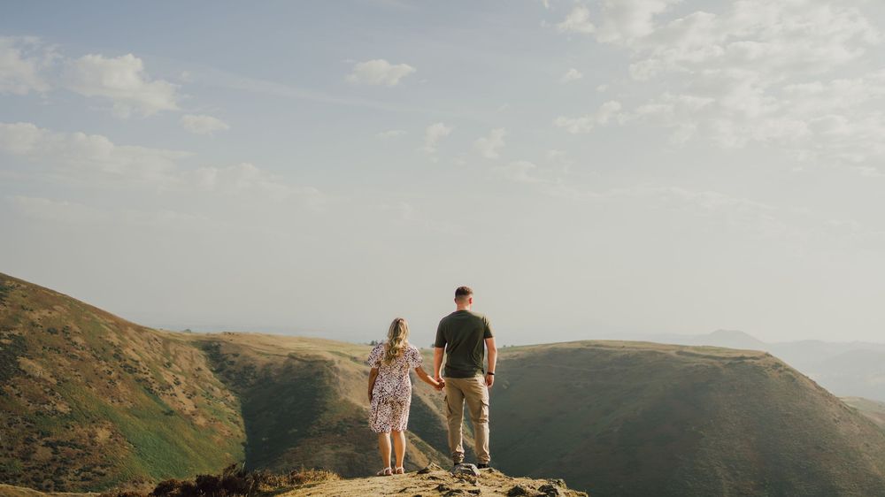 Two people standing together on a scenic mountain overlook during golden hour with rolling hills in the background.