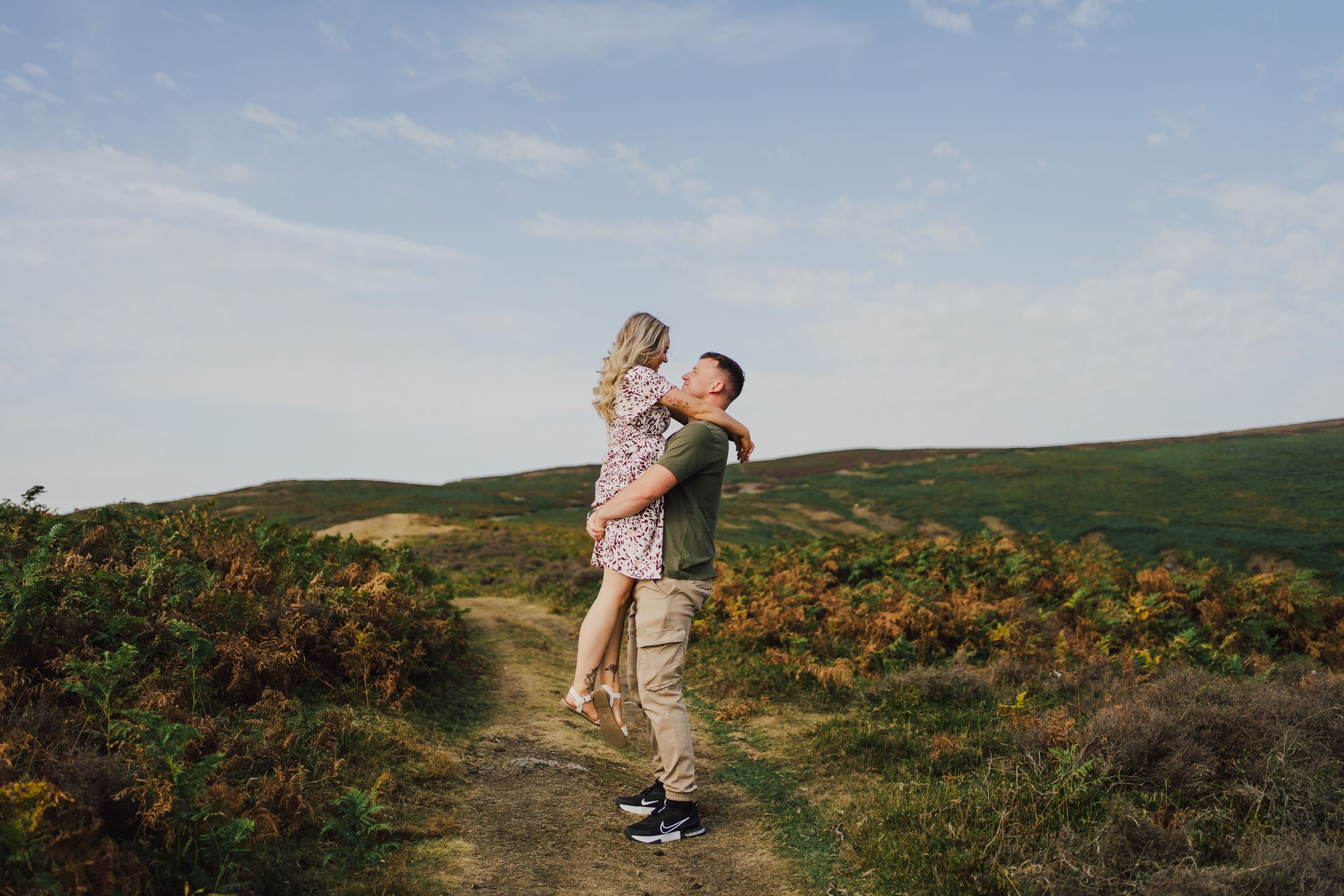 Two people sharing a joyful moment on a scenic hillside trail surrounded by yellow wildflowers and green slopes.