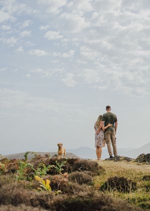 Couple standing with their dog on a scenic hilltop overlooking mountain landscape on a cloudy summer day.
