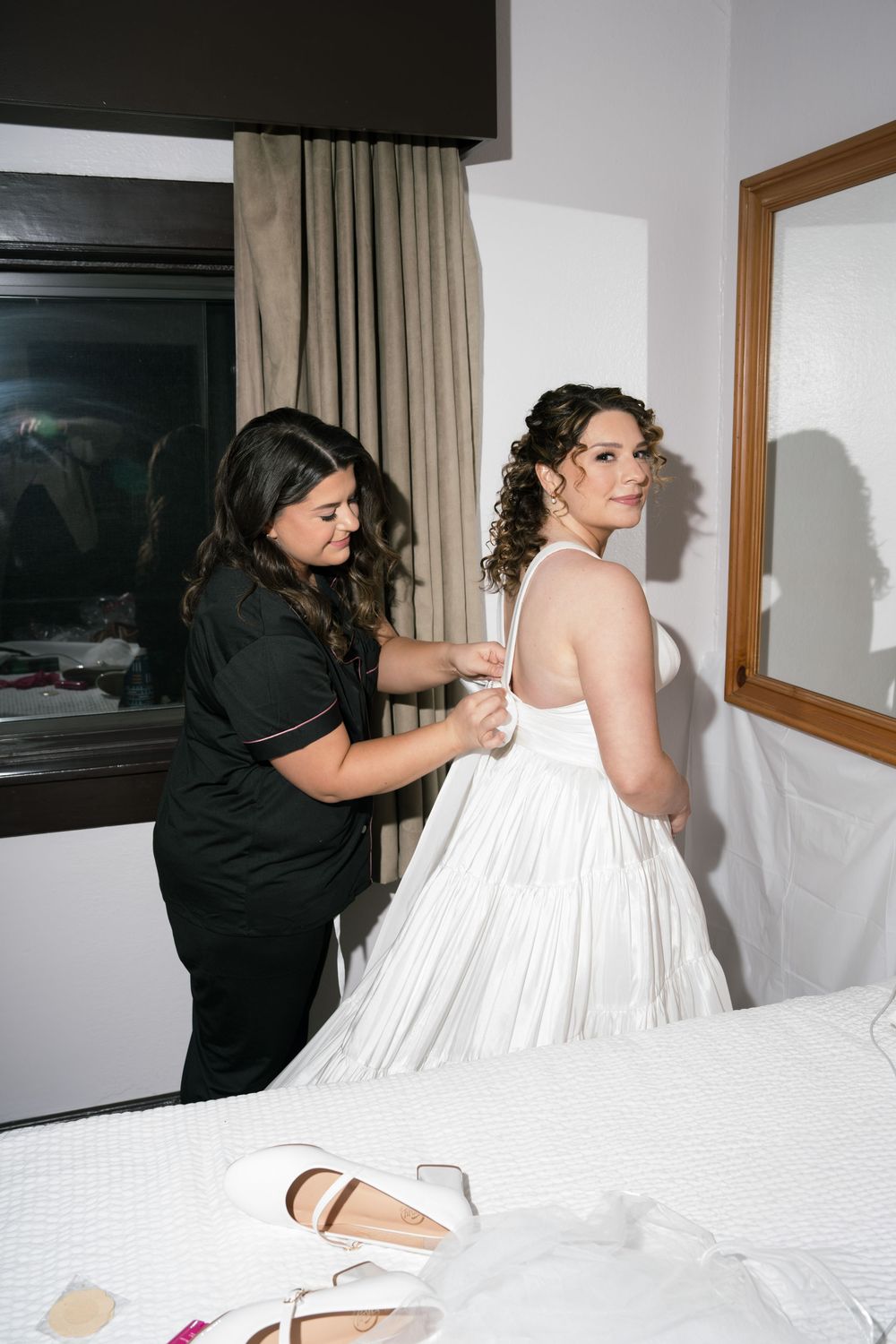 A bridesmaid in black helps a bride in a white gown get dressed in a hotel room before the wedding ceremony.