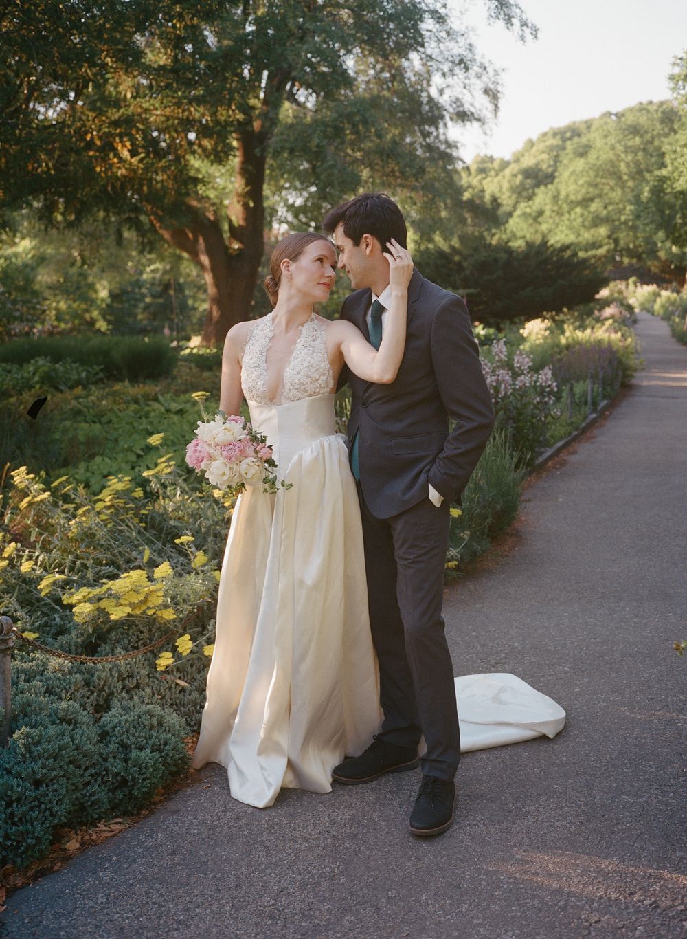 Bride in champagne gown holding pink bouquet leans close to groom in dark suit on garden path.