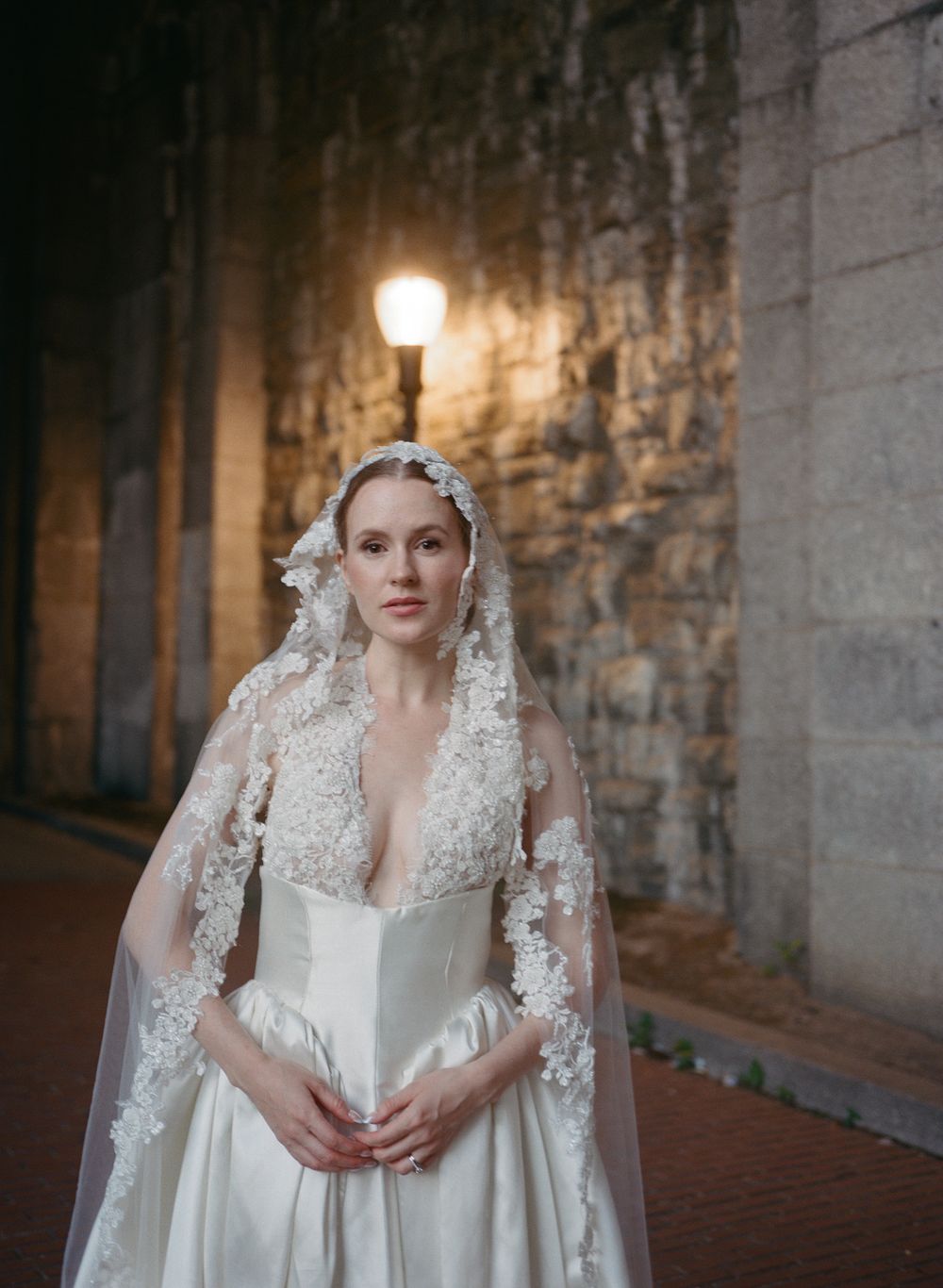 Bride in a deep V-neck wedding gown and floral lace veil poses under a glowing street lamp against a stone wall.