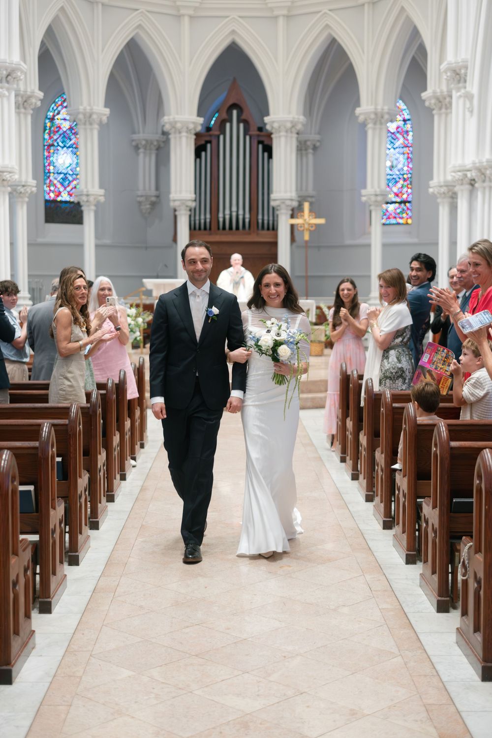 Newlyweds walk down church aisle after ceremony, bride in white gown holds bouquet as guests applaud in elegant cathedral.
