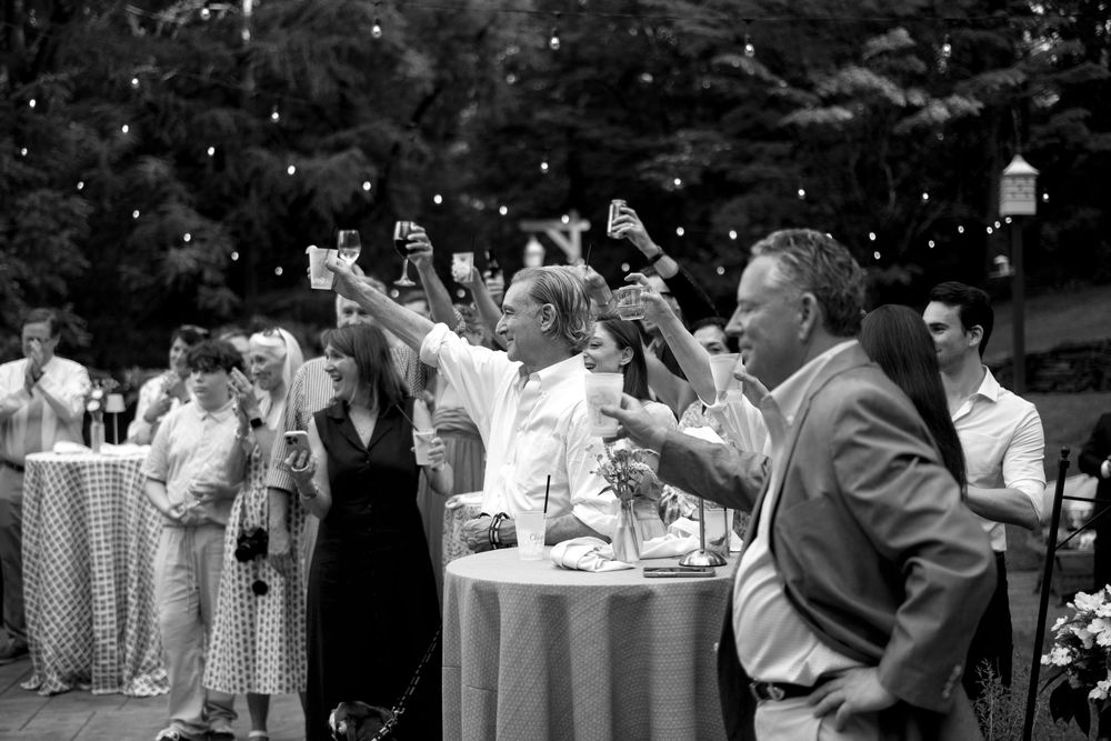 Black and white photo of wedding guests raising glasses in a toast at an outdoor reception with string lights.