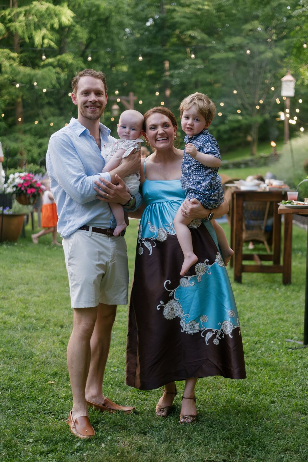 A smiling family of four poses outdoors at a garden party, parents holding two young children amid string lights.