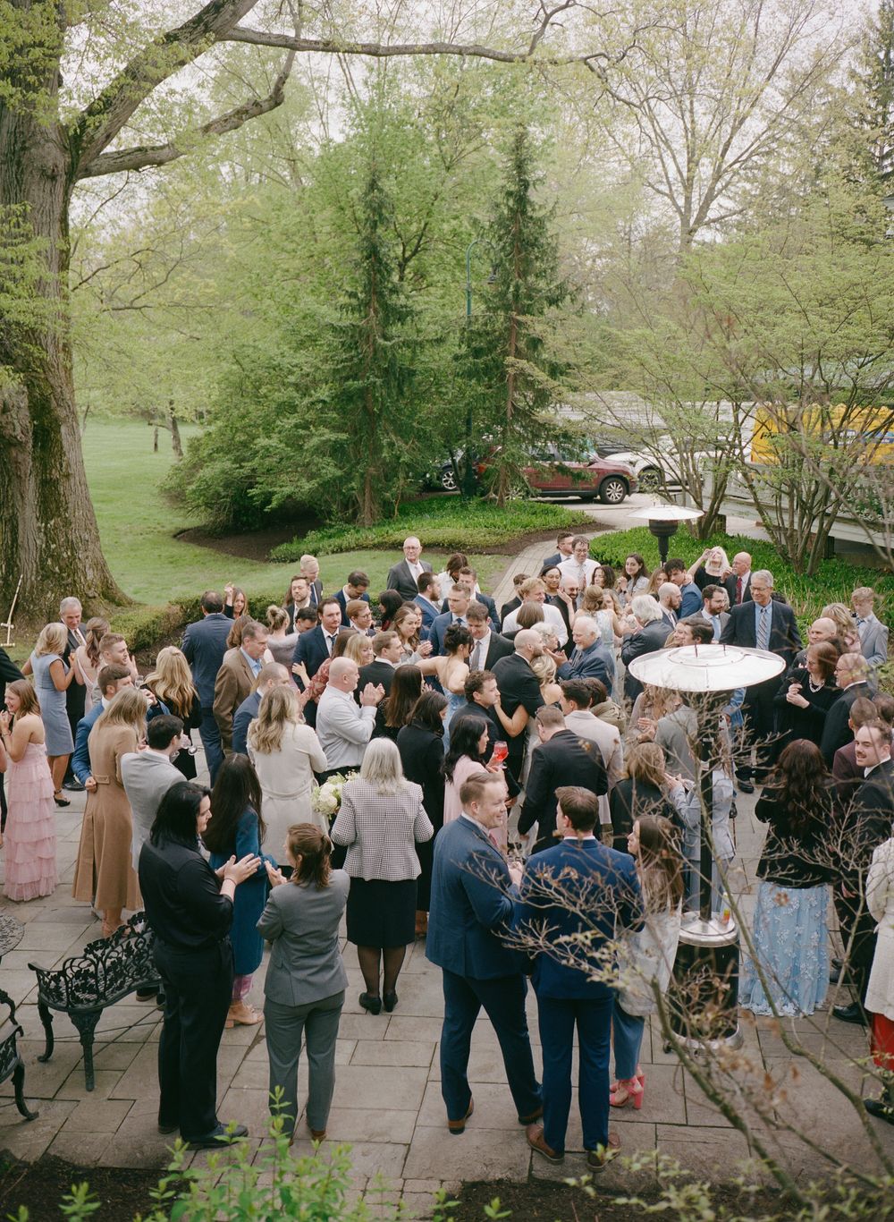 Aerial view of elegant outdoor wedding cocktail hour on stone patio surrounded by lush spring trees.