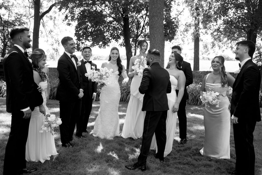Black and white wedding party photo with bride, groom, and guests gathered outdoors under trees.