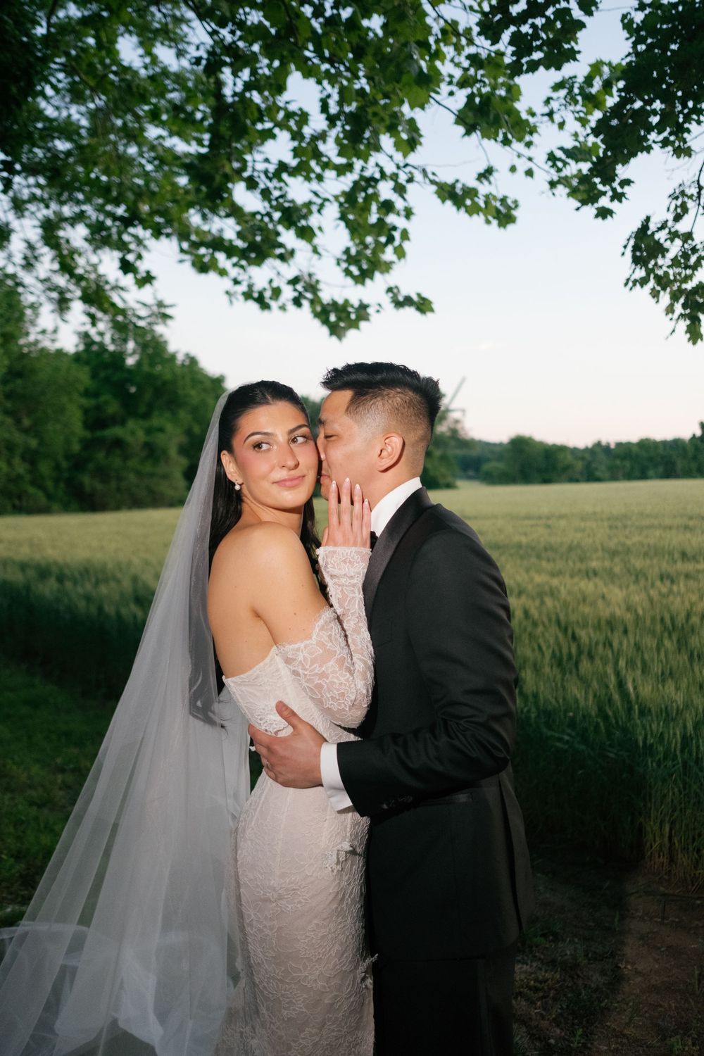 Groom in black tuxedo kissing bride in lace off-shoulder gown and veil beside a green field at sunset.