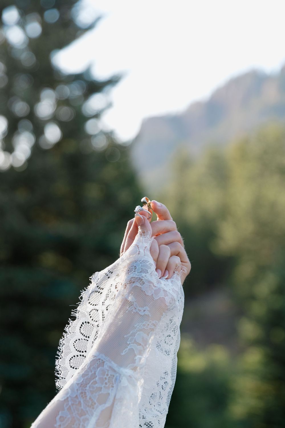 A bride in a white lace sleeve raises her hand holding a small flower with green trees and mountains in the background.