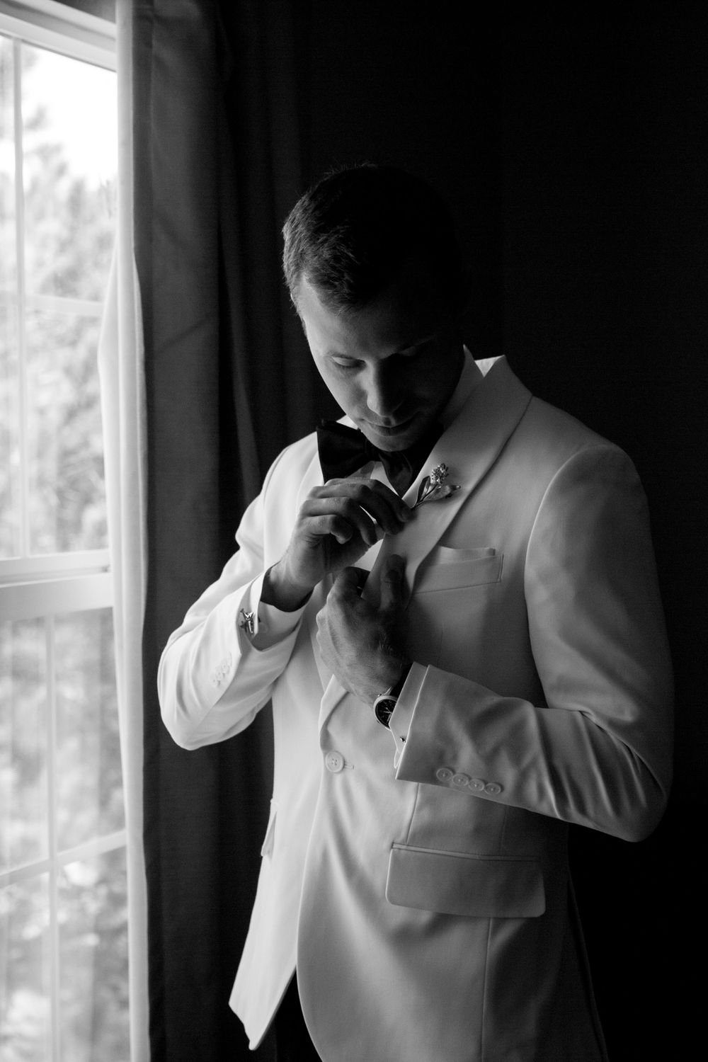 Groom in white tuxedo jacket adjusting bow tie by window in elegant black and white wedding portrait.