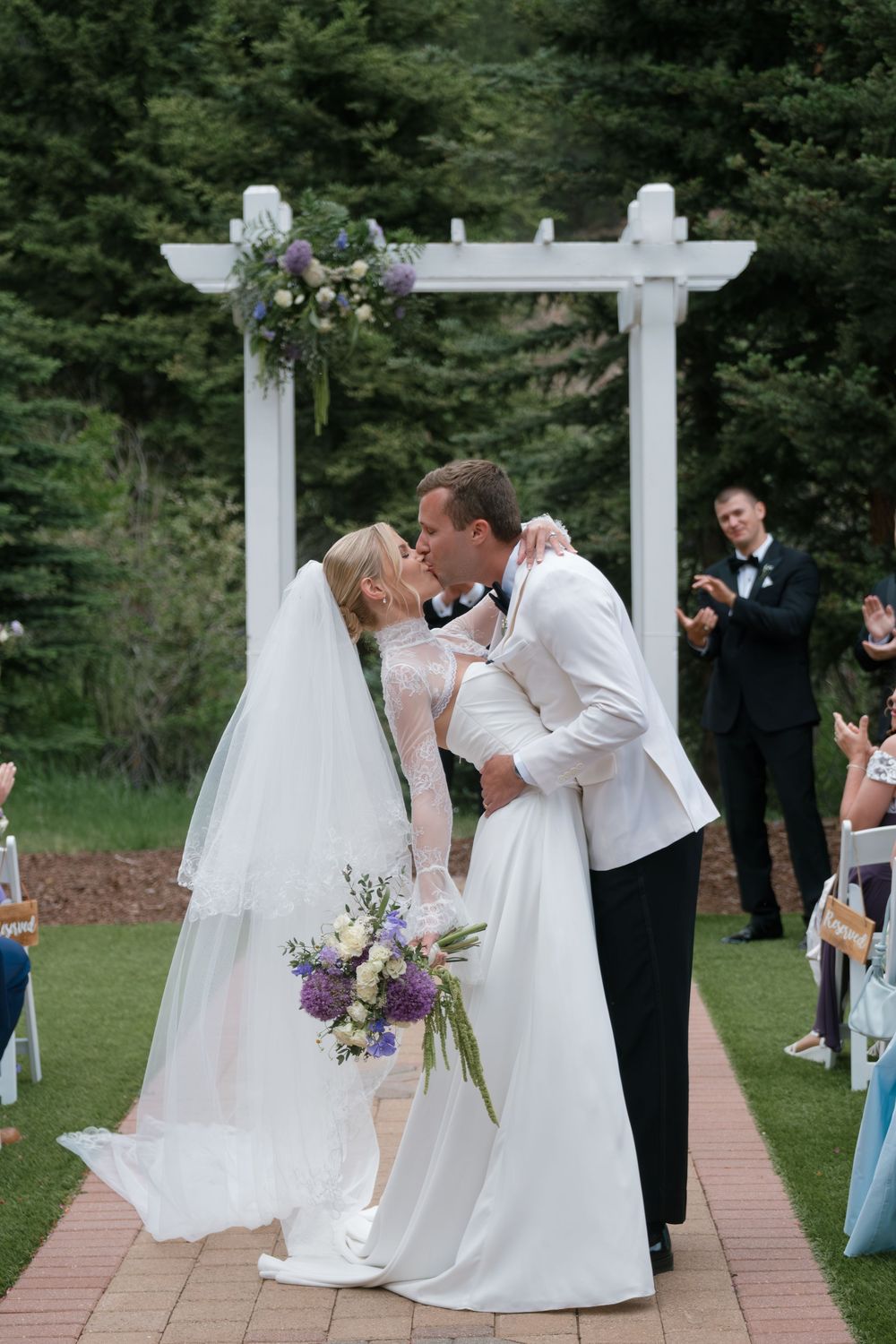 Bride and groom share first kiss at outdoor wedding ceremony under white pergola with purple floral arrangement.
