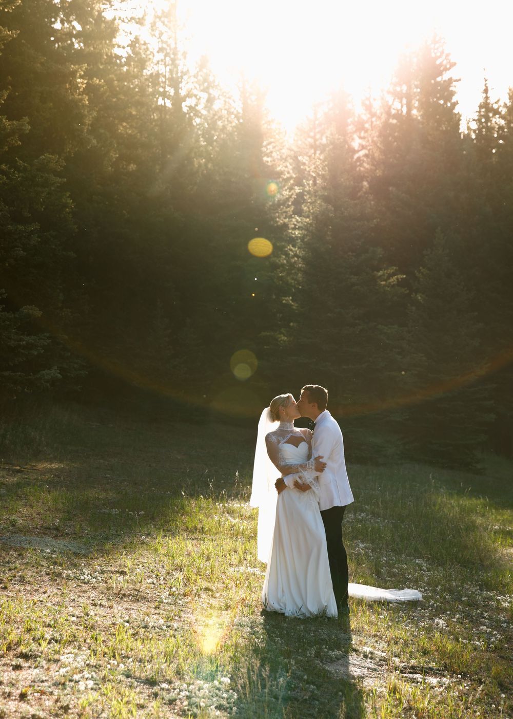 Bride and groom sharing a kiss in a sunlit forest clearing, golden hour light streaming through tall trees.