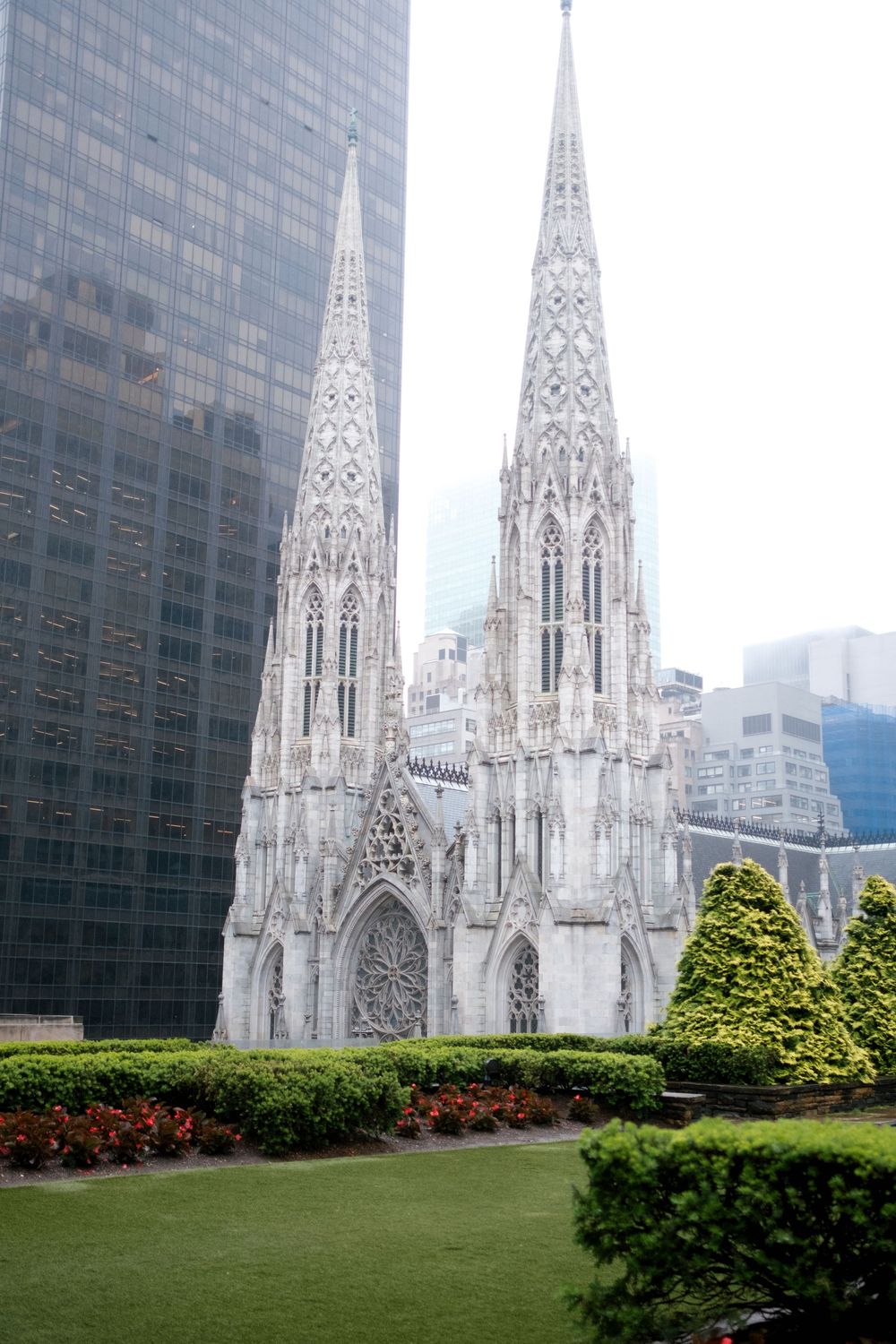St. Patrick's Cathedral Gothic spires rise between modern skyscrapers in Midtown Manhattan, surrounded by manicured gardens.