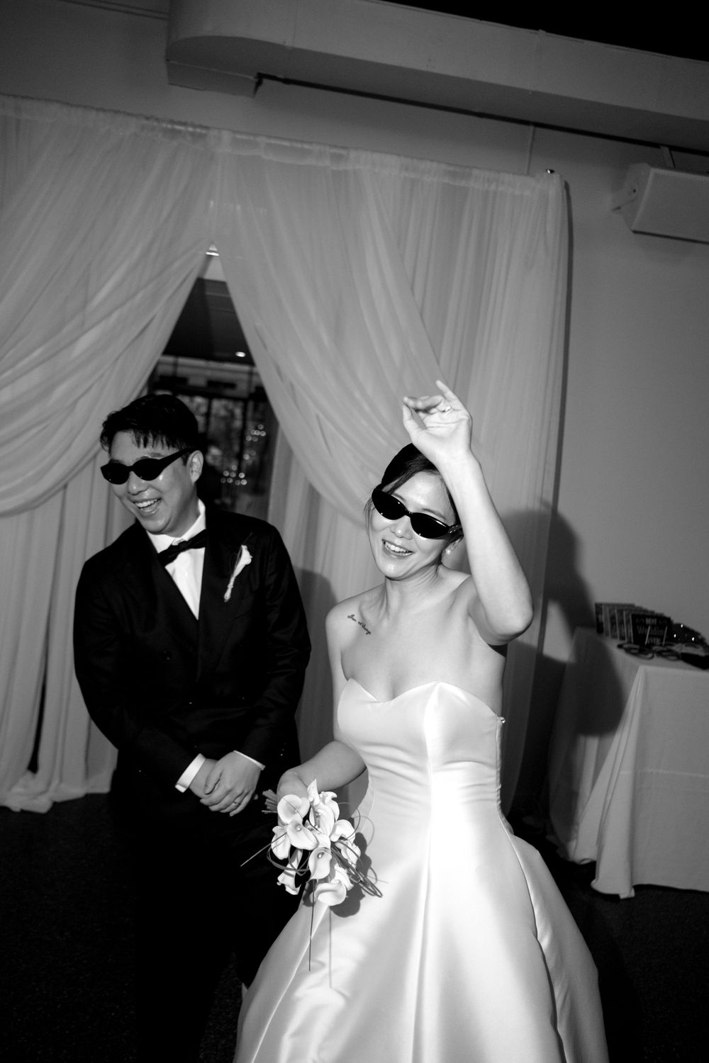 A joyful bride and groom wearing sunglasses laugh together at their wedding reception in black and white photo.
