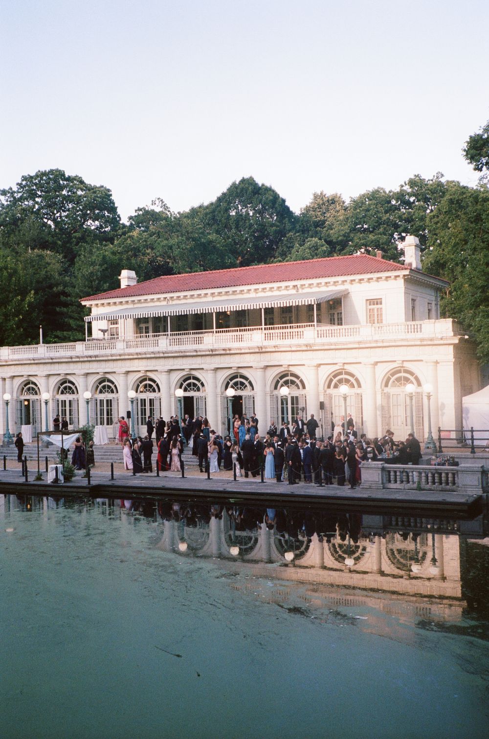 Elegant waterfront boathouse venue with guests gathered on terrace for outdoor event, reflecting in calm water below.