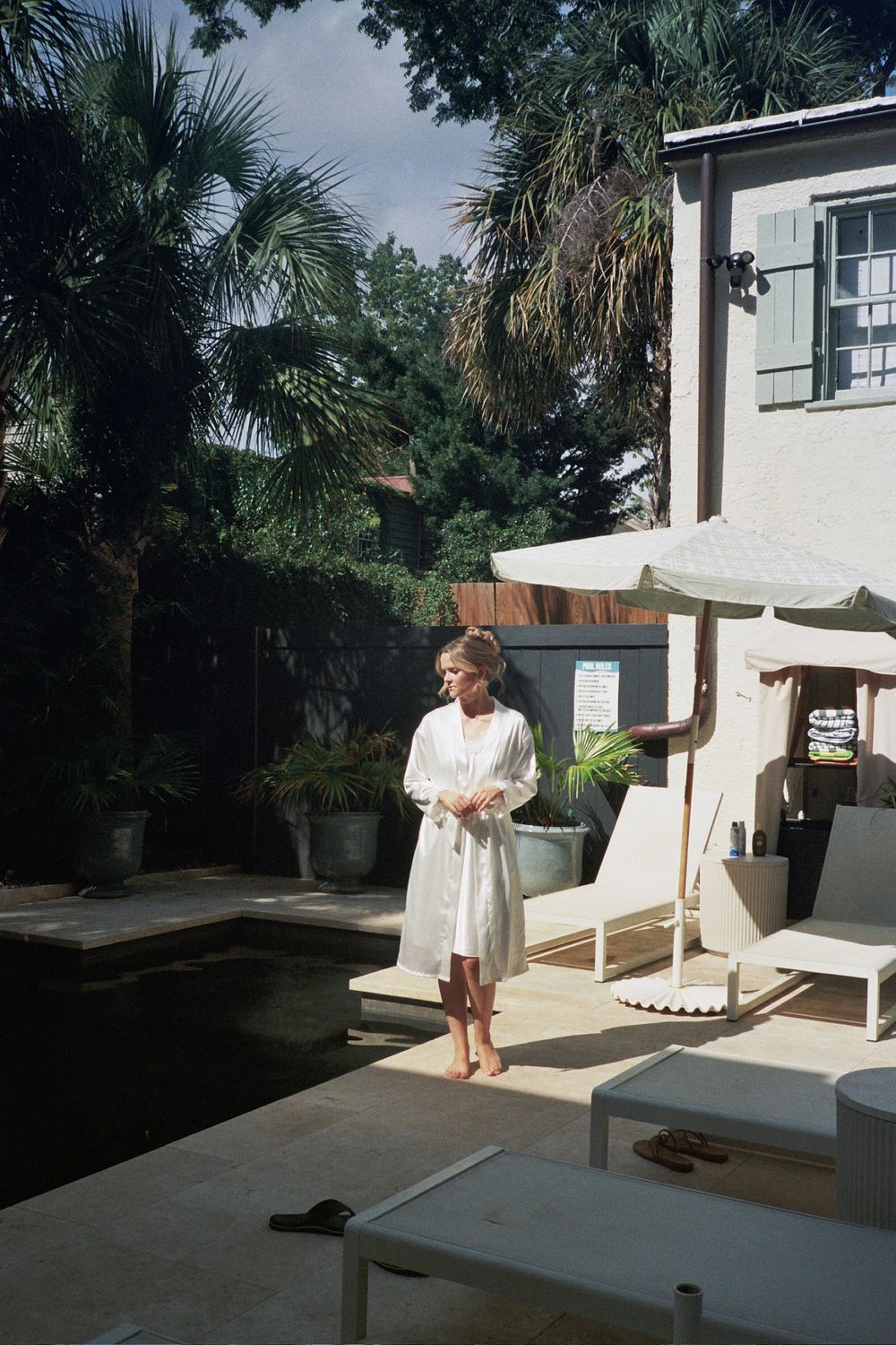 Woman in white robe standing barefoot by a serene outdoor pool surrounded by lush palm trees and elegant patio furniture.
