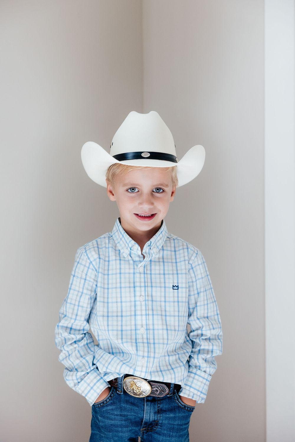 Young child in western attire wearing plaid button-down shirt, jeans and white cowboy hat against neutral background.