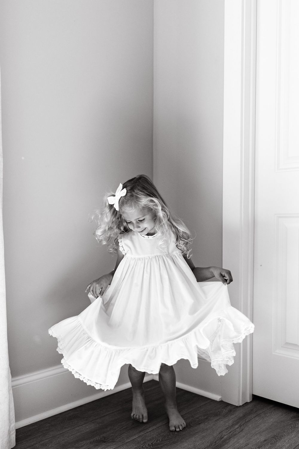 A black and white artistic photograph shows someone in a white dress peeking around a corner in a minimalist setting.