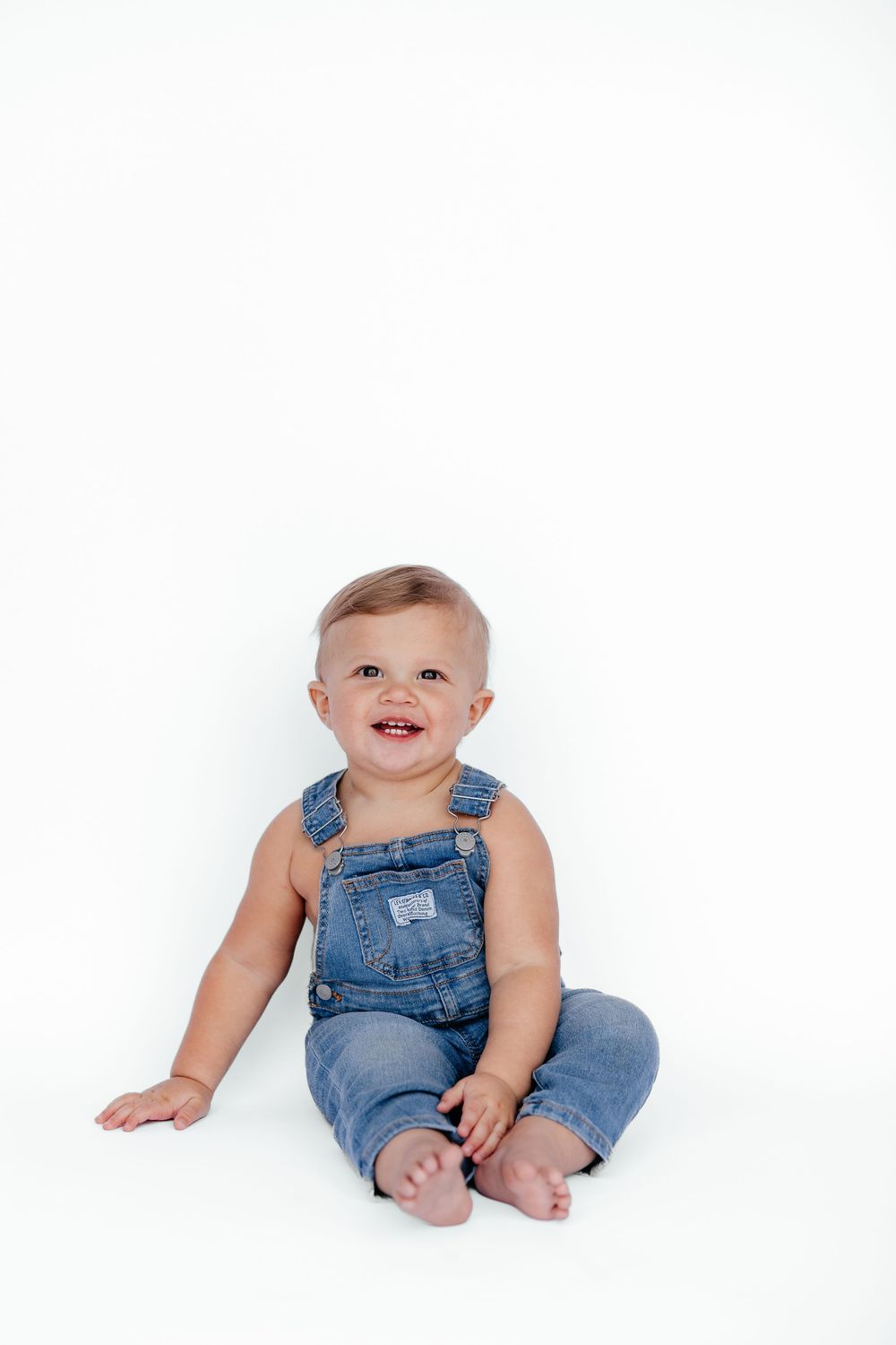 Baby wearing denim overalls sits on white background in four different playful poses.