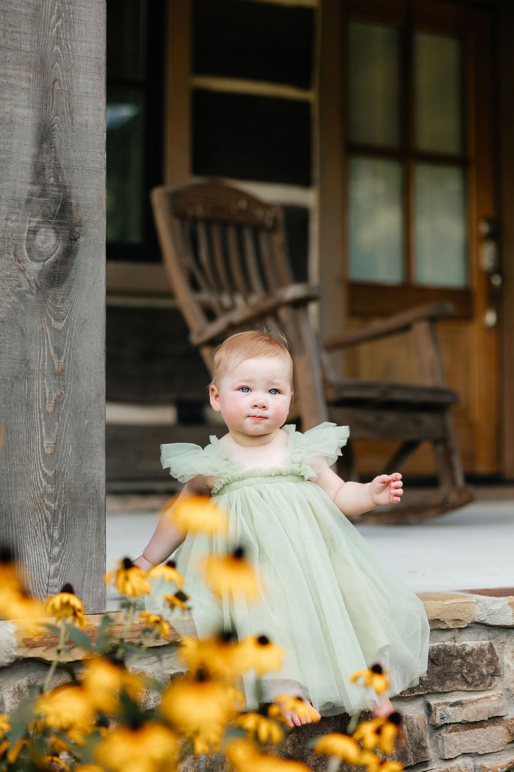 Baby in mint green dress sits on rustic porch with yellow black-eyed susan flowers blooming in foreground.