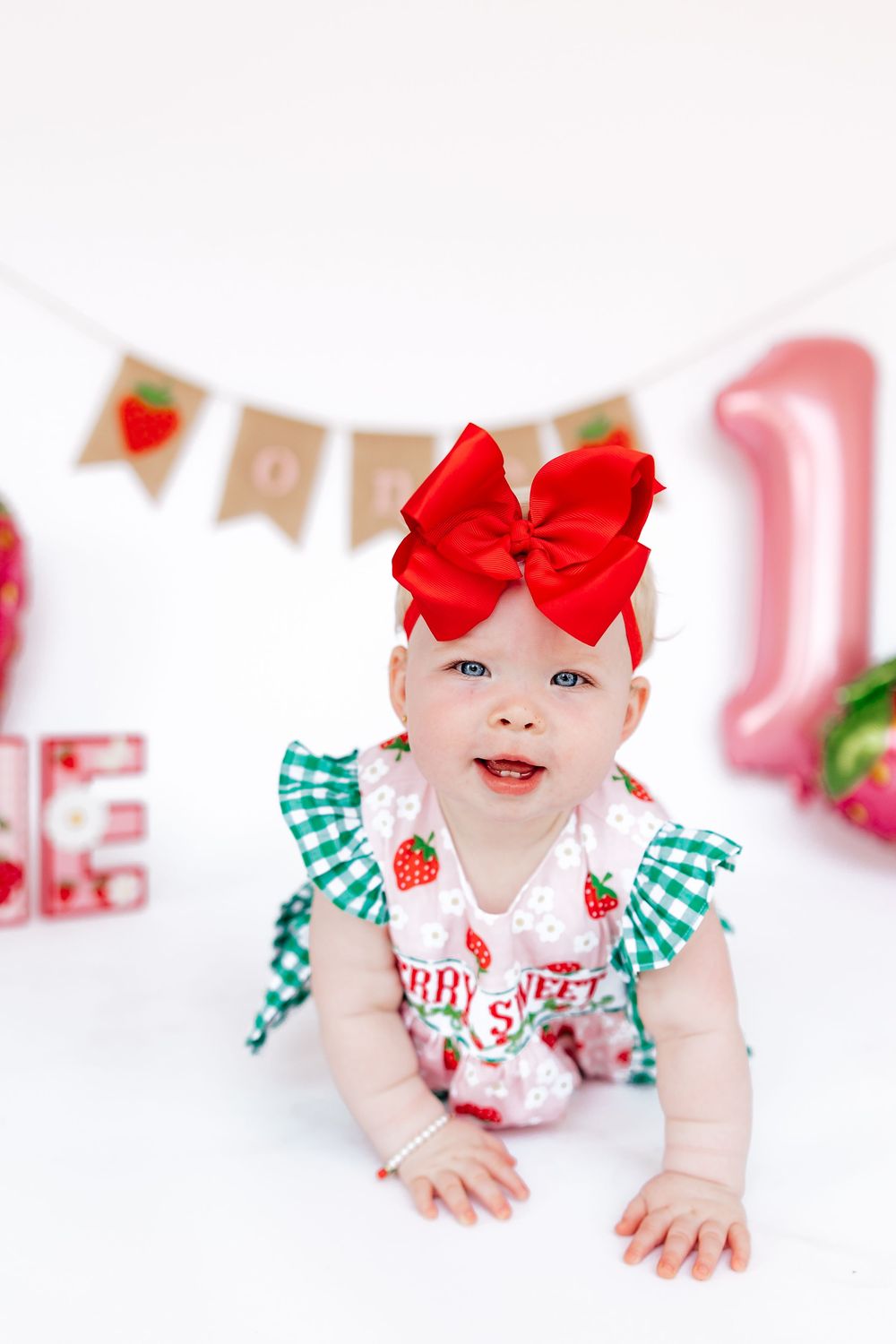 Baby wearing green gingham and strawberry print outfit with red bow crawling on white background.