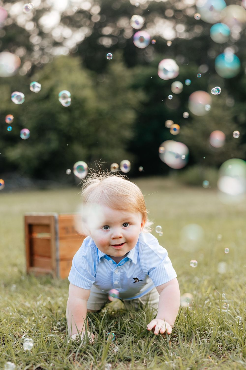 A happy baby boy in a blue shirt crawls on green grass surrounded by floating soap bubbles outdoors.