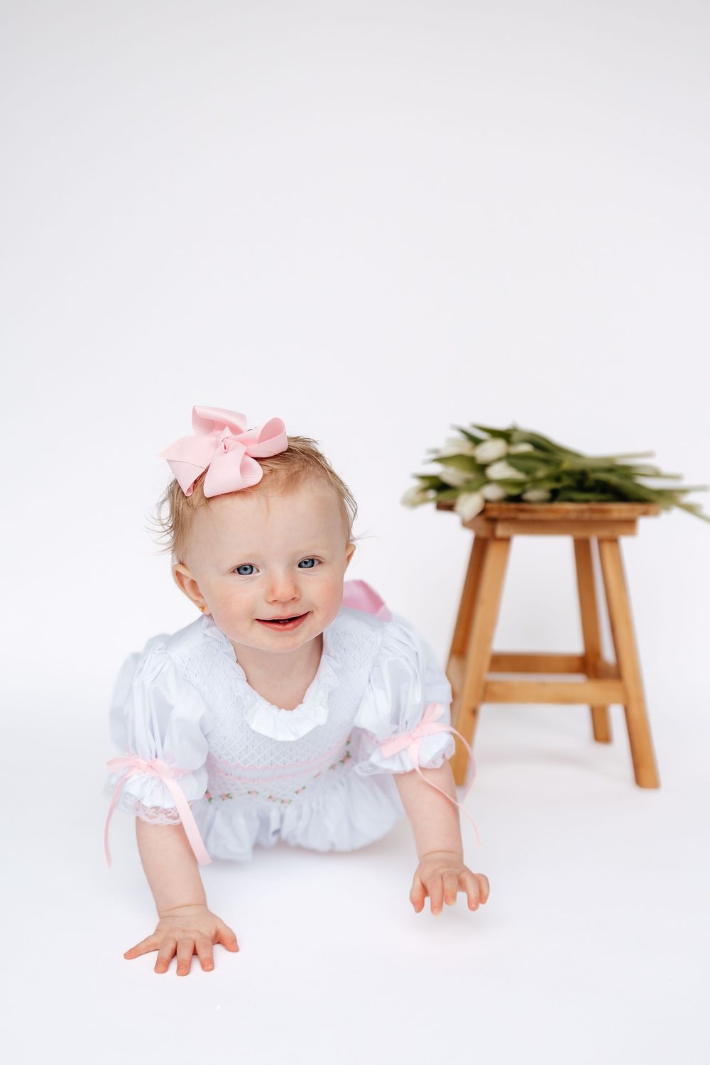 A smiling baby girl in a white and pink dress with a bow headband crawls on a white floor near a wooden stool with tulips.