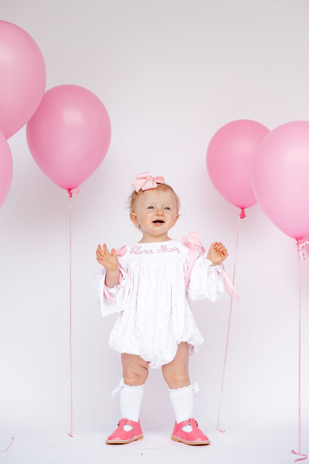 Smiling baby girl in white dress with pink bow, knee socks, and pink shoes surrounded by pink balloons.