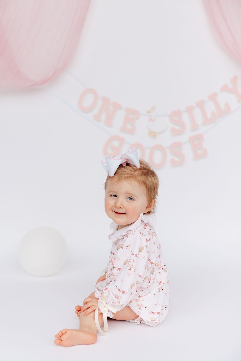 Smiling baby girl in floral romper with white bow sits before a pink One Silly Goose banner on white backdrop.