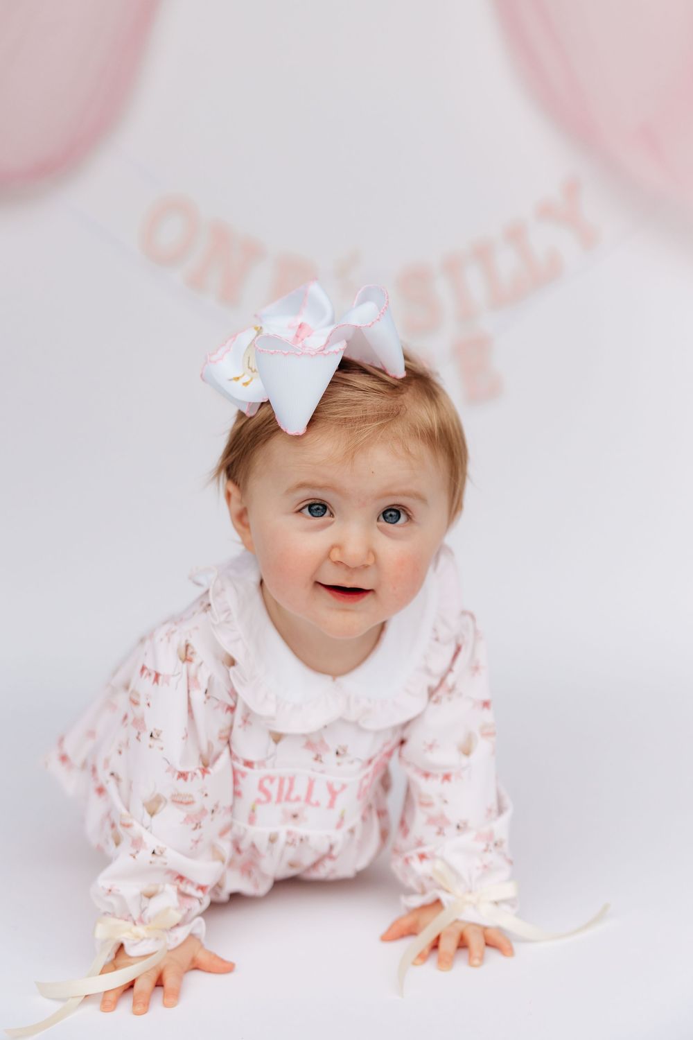 Adorable baby girl in pink floral romper with white bow headband crawling at a first birthday photo shoot.
