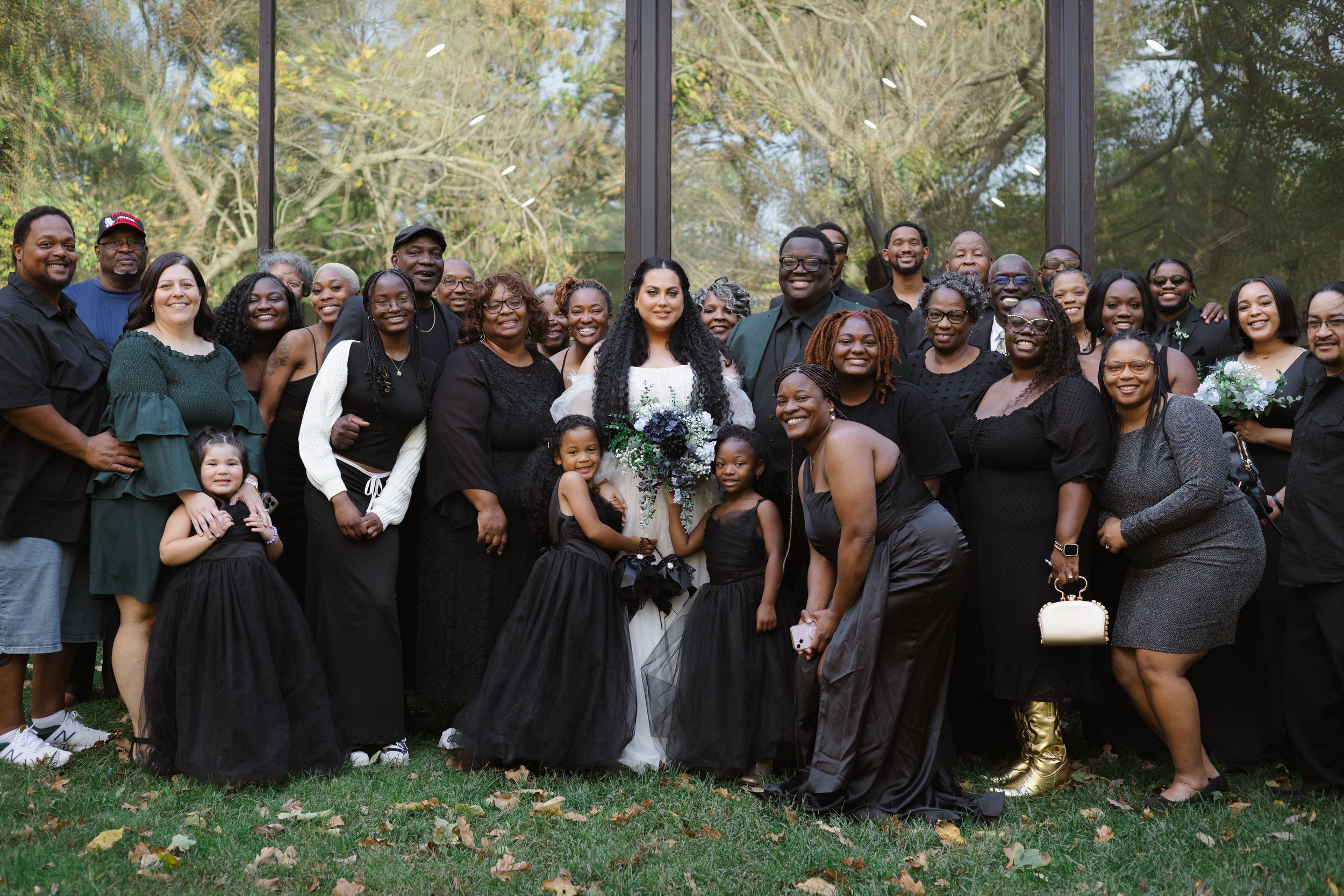 A wedding party poses outdoors in coordinated black attire for a celebratory group photo.