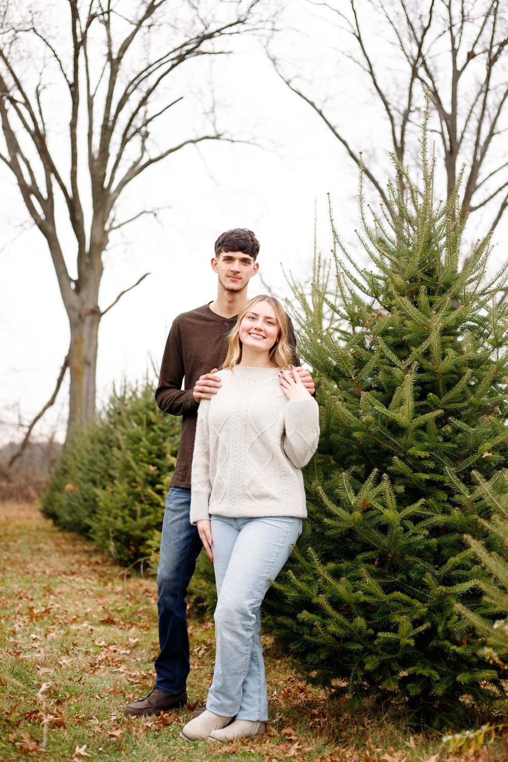 Young couple posing together at a Christmas tree farm, surrounded by evergreen trees on a cloudy winter day.