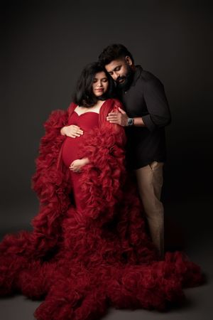 Dramatic maternity photoshoot with woman in flowing red tulle dress against dark background.
