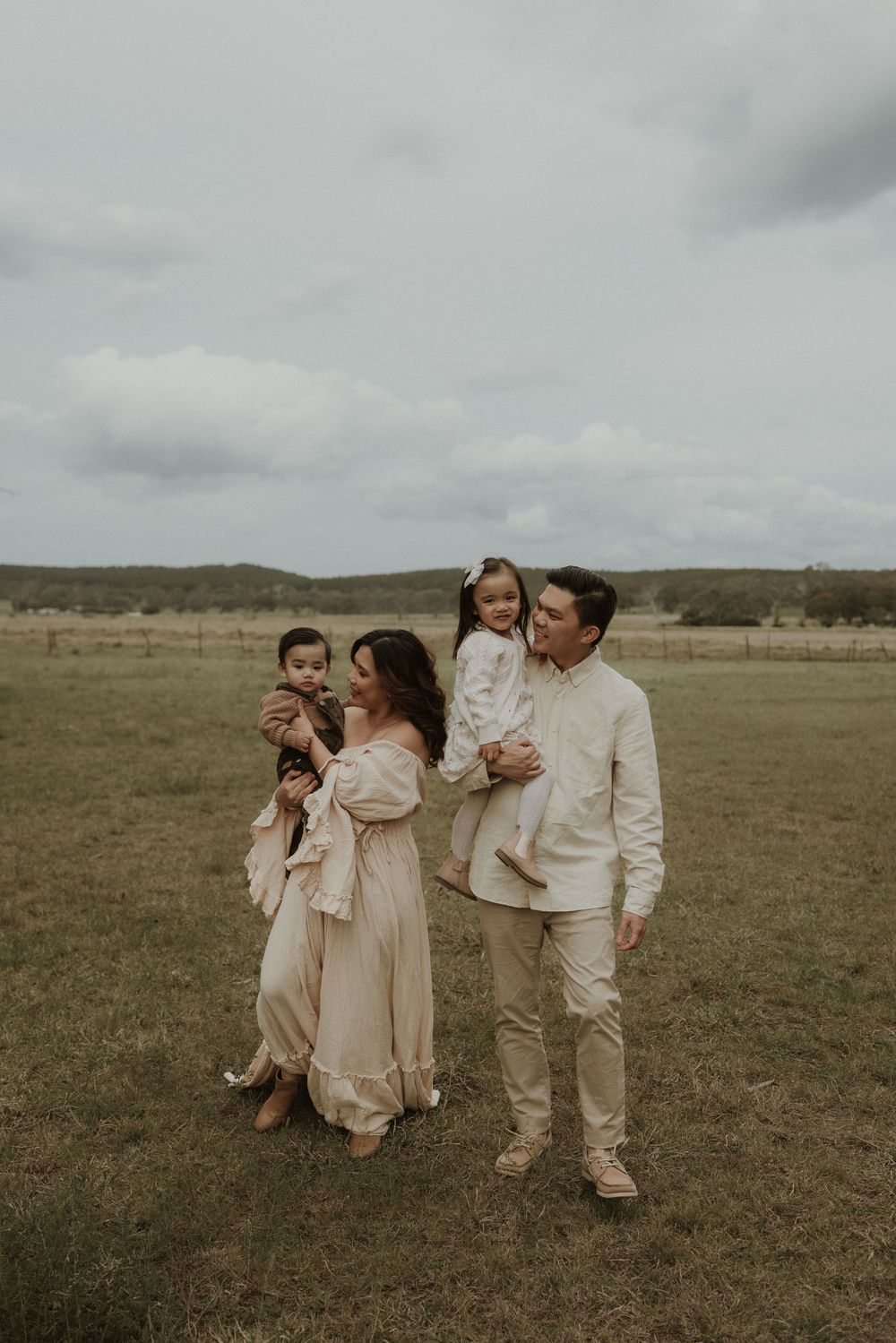 A family in neutral-colored outfits shares tender moments together while standing in an open field under cloudy skies.