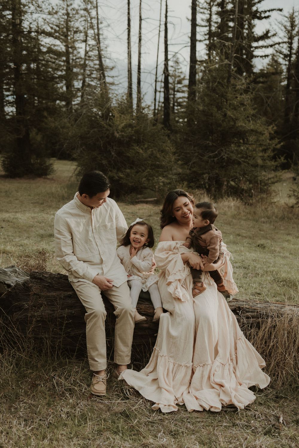 A family wearing coordinated cream-colored outfits poses together in a scenic forest setting at sunset.