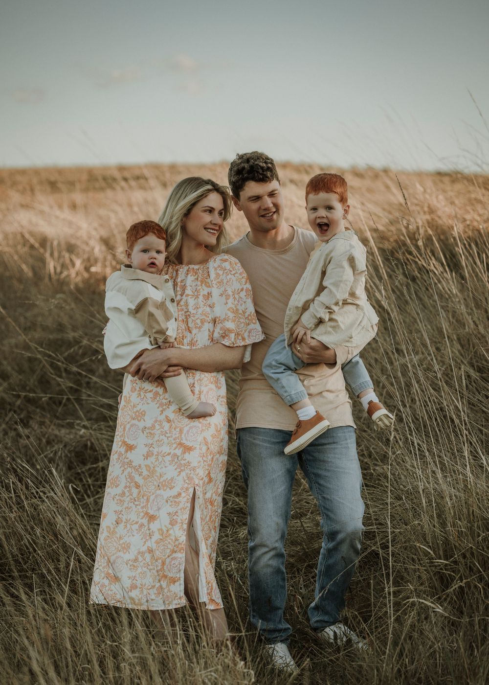 A family stands together in a field of tall grass at sunset wearing casual clothing.