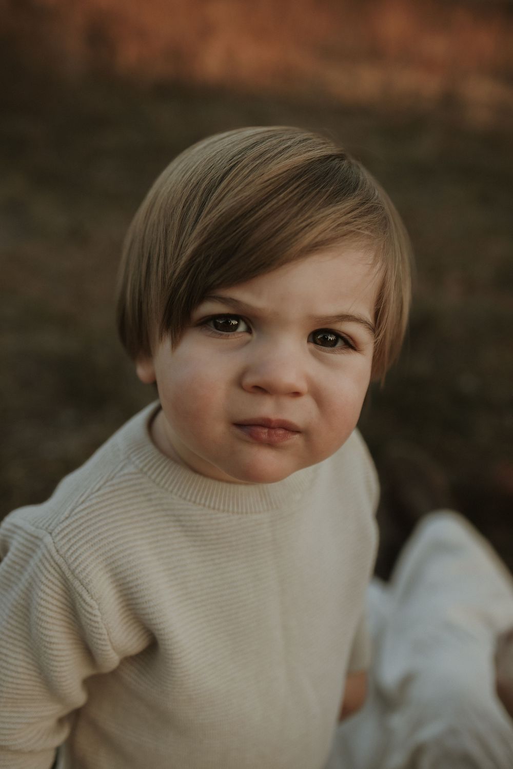 A young child in a cream colored sweater gazes at the camera with a slight smile during a warm outdoor portrait session.