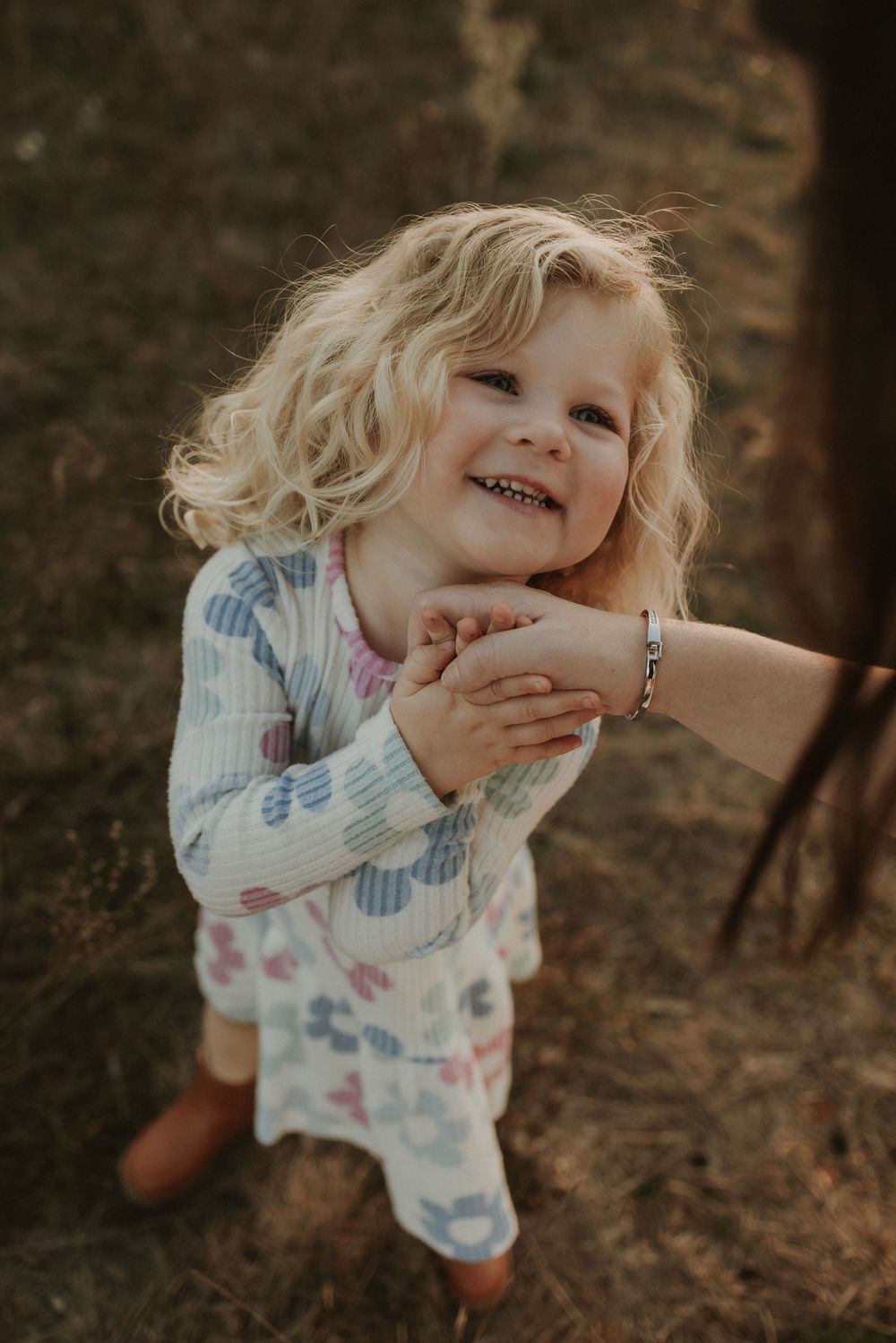 A young child in floral pajamas smiles while holding an adult's hand outdoors in warm lighting.