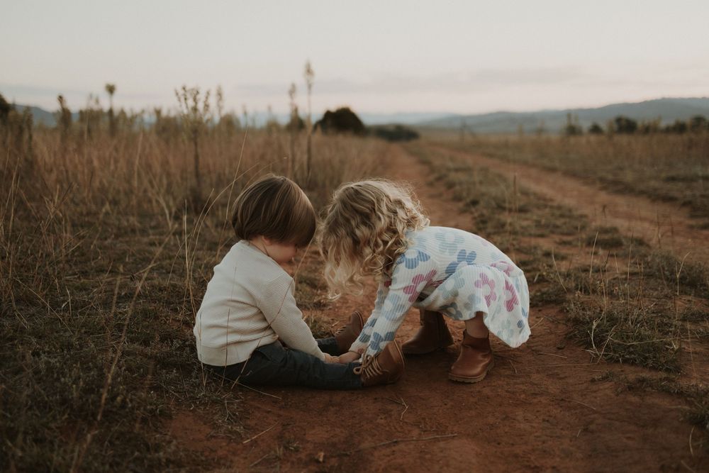 Two children in casual wear crouch together on a dirt path at sunset examining something on the ground.