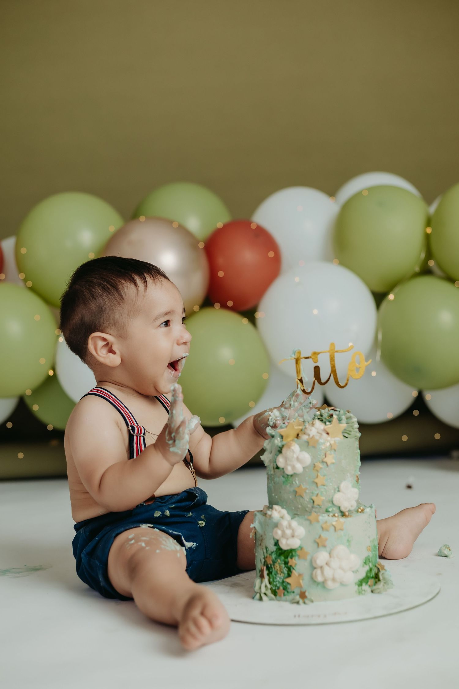 A joyful cake smash photo with green and white balloons and a two-tier birthday cake.